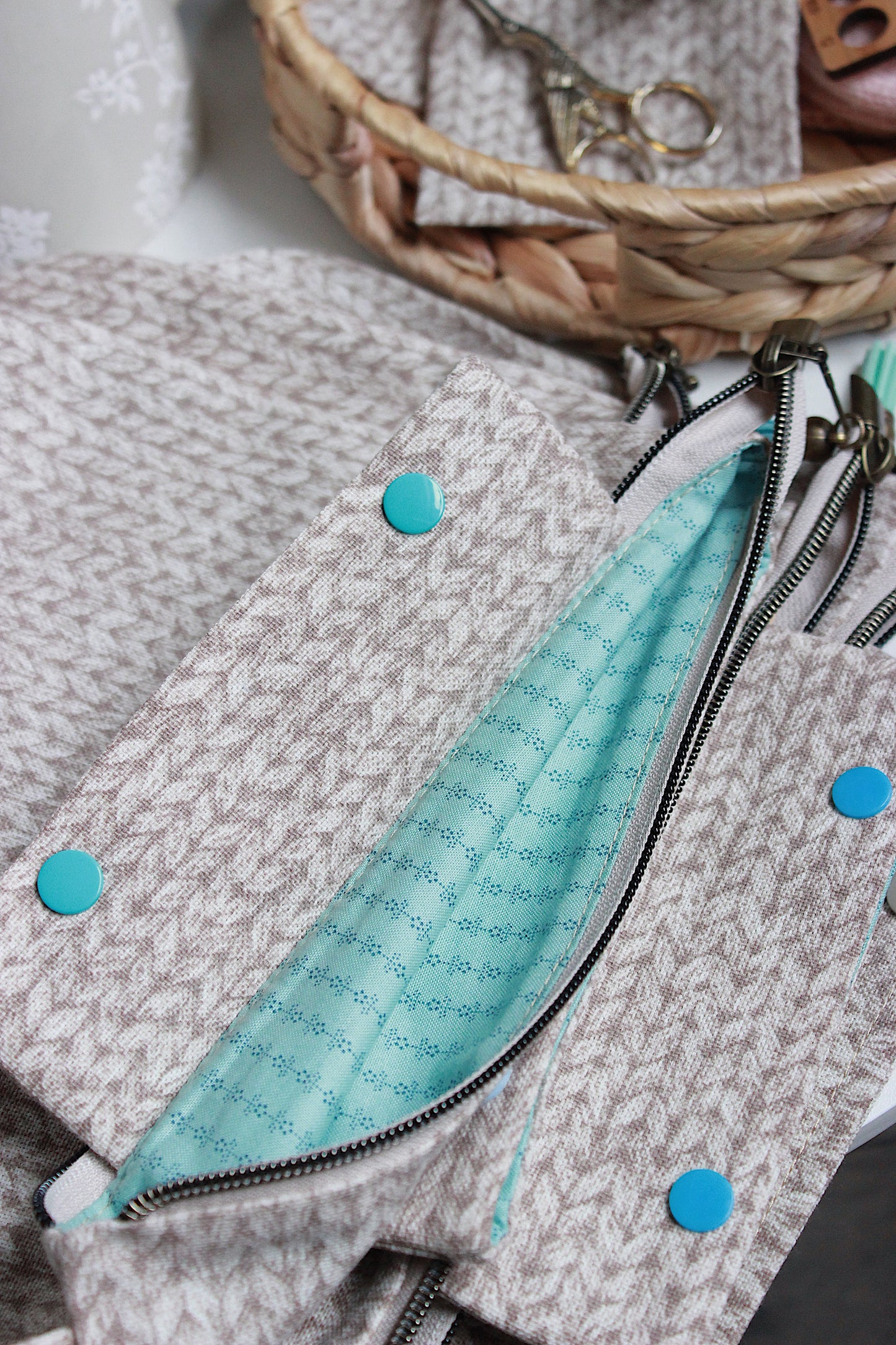 Close-up of a beige knitting project bag with a turquoise interior, showcasing zippers and button details.